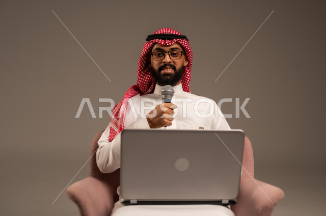 Conducting interviews and asking questions, using modern technology devices, close-up portrait of a Saudi Arabian Gulf man wearing traditional thobe and shemagh sitting on a chair using a laptop and holding a loudspeaker looking at the camera and giving speeches, gray background
