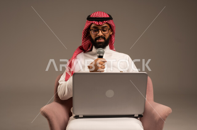 إجراء المقابلات وطرح الأسئلة، استخدام الأجهزة التقنية الحديثة، بورتريه مقرب لرجل عربي خليجي سعودي يرتدي الثوب التقليدي والشماغ يجلس على الكرسي يستخدم الحاسوب المحمول ويحمل بيده مكبر الصوت يقوم بإلقاء الخطابات، خلفية رمادية