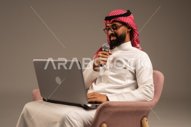 Conducting interviews and asking questions, using modern technology devices, close-up portrait of a Saudi Arabian Gulf man wearing traditional thobe and shemagh sitting on a chair using a laptop and holding a loudspeaker giving speeches, gray background
