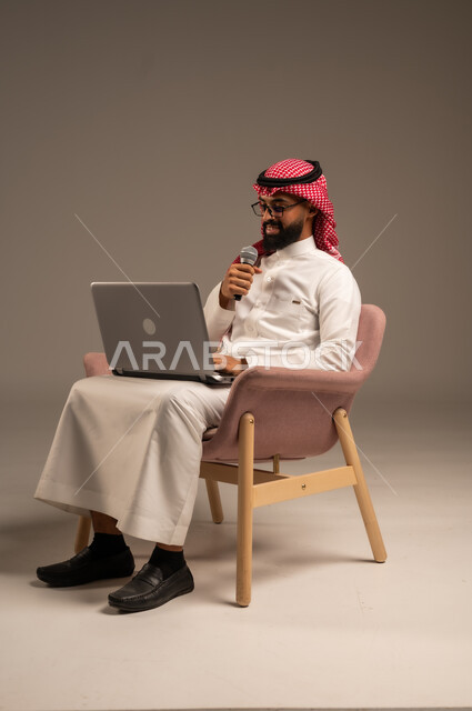 Conducting interviews and asking questions, using modern technical devices, portrait of a Saudi Arabian Gulf man wearing traditional thobe and shemagh sitting on a chair using a laptop and holding a loudspeaker giving speeches, gray background