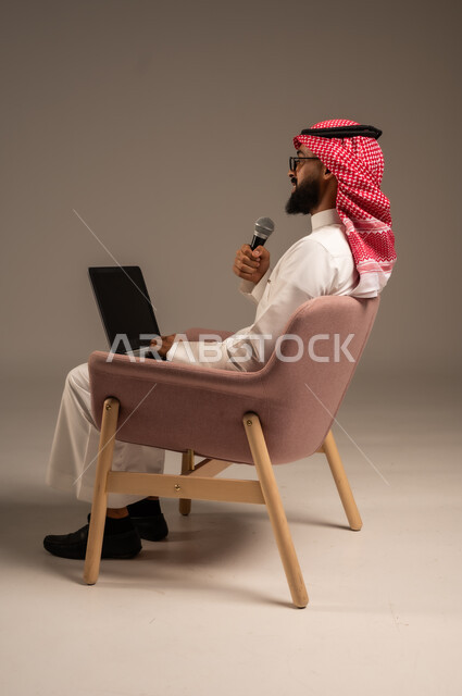 استخدام الأجهزة التقنية الحديثة، بورتريه من الجانب لرجل عربي خليجي سعودي يرتدي الثوب التقليدي والشماغ يجلس على الكرسي يستخدم الحاسوب المحمول ويحمل بيده مكبر الصوت يقوم بإلقاء الخطابات، إجراء المقابلات وطرح الأسئلة، خلفية رمادية