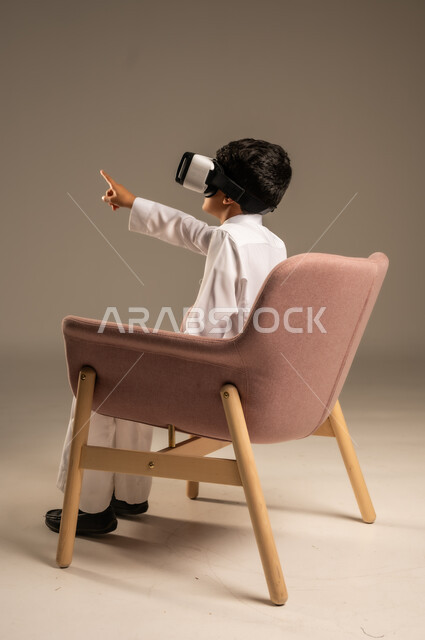 استخدام حاسة اللمس لملامسة عنصر ما بإيماءات النقر، تقنيات حديثة متطورة، بورتريه لولد عربي خليجي سعودي يرتدي الثوب التقليدي ونظارة الواقع الافتراضي ثلاثية الأبعاد يجلس على كرسي مريح  ويشير إلى شيء ما بأصبع السبابة، محاكاة العالم الخيالي، خلفية رمادية