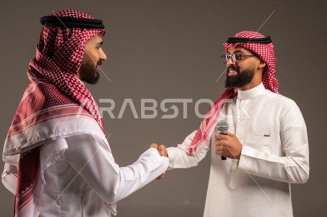 التواصل الفعال وتبادل الحوارات والنقاشات، إجراء المقابلات وطرح الأسئلة، بورتريه مقرب لرجل عربي خليجي سعودي يرتدي الثوب التقليدي والشماغ يحمل بيده مكبر الصوت يصافح رجل آخر بتعابير الترحيب والاستقبال، خلفية رمادية