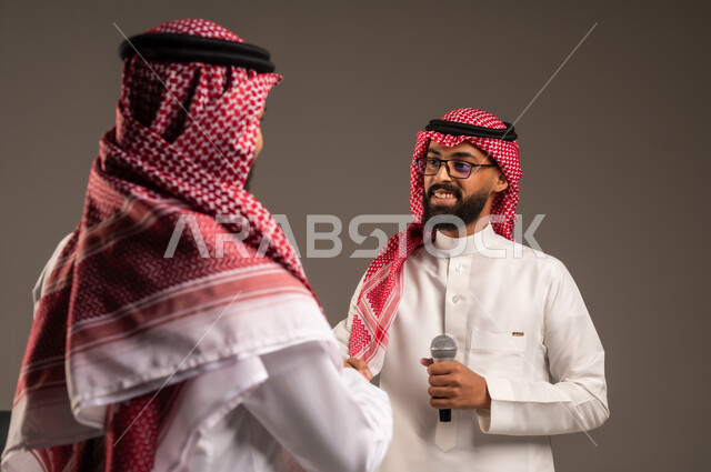 التواصل الفعال وتبادل الحوارات والنقاشات، إجراء المقابلات وطرح الأسئلة، بورتريه مقرب لرجل عربي خليجي سعودي يرتدي الثوب التقليدي والشماغ يحمل بيده مكبر الصوت يصافح رجل آخر بتعابير الترحيب والاستقبال، خلفية رمادية