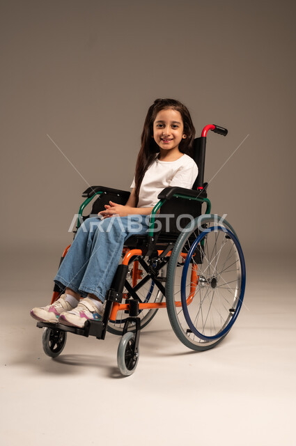 ضعف القدرة الحركية وصعوبة المشي، بورتريه لفتاة صغيرة عربية خليجية سعودية من ذوي الاحتياجات الخاصة ترتدي الزي الكاجوال تجلس على كرسي متحرك وتنظر للكاميرا بإيماءات البشاشة والسرور، الجسم بالطول الكامل، خلفية رمادية