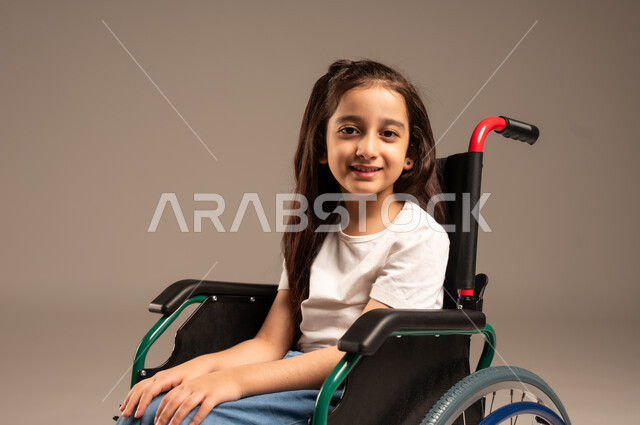Poor motor ability and difficulty walking, close-up portrait of a young Saudi Arabian Gulf girl with special needs wearing casual clothes sitting on a wheelchair and looking at the camera with gestures of cheerfulness and pleasure, gray background