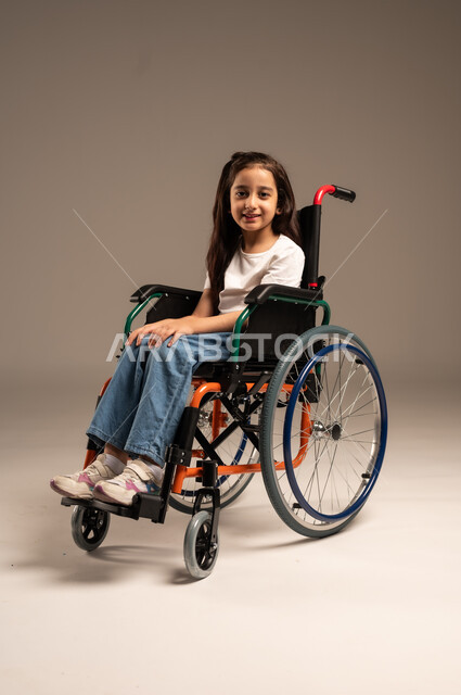 ضعف القدرة الحركية وصعوبة المشي، بورتريه لفتاة صغيرة عربية خليجية سعودية من ذوي الاحتياجات الخاصة ترتدي الزي الكاجوال تجلس على كرسي متحرك وتنظر للكاميرا بإيماءات البشاشة والسرور، الجسم بالطول الكامل، خلفية رمادية