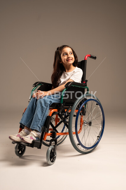 والنظرإلى الأعلى بإيماءات الأمل والتفاؤل، بورتريه لفتاة صغيرة عربية خليجية سعودية من ذوي الاحتياجات الخاصة ترتدي الزي الكاجوال تجلس على كرسي متحرك تعاني من ضعف القدرة الحركية وصعوبة في المشي، الجسم بالطول الكامل، خلفية رمادية