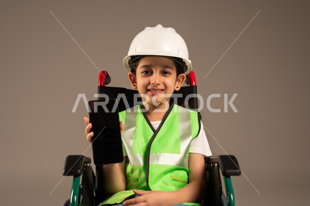 عرض شاشة هاتف محمول سوداء فارغة، تجسيد العمل في المجال الهندسي منذ الصغر، بورتريه مقرب لولد عربي خليجي سعودي من ذوي الاحتياجات الخاصة يرتدي قبعة وسترة الحماية يجلس على كرسي متحرك يمسك جهاز الجوال بيده وينظر للكاميرا بإيماءات السرور، خلفية بيج