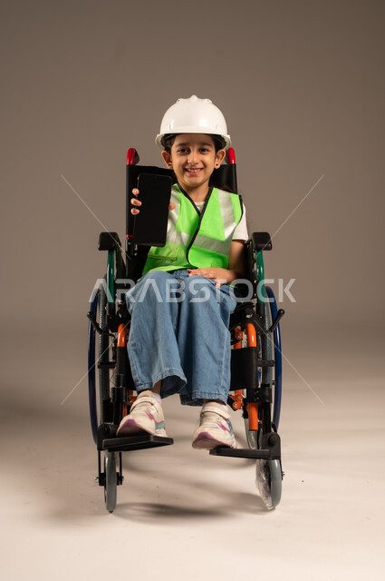 عرض شاشة هاتف محمول سوداء فارغة، تجسيد العمل في المجال الهندسي منذ الصغر، بورتريه لولد عربي خليجي سعودي من ذوي الاحتياجات الخاصة يرتدي قبعة وسترة الحماية يجلس على كرسي متحرك يمسك جهاز الجوال بيده وينظر للكاميرا بإيماءات السرور، الجسم بالطول الكامل، خلفية بيج