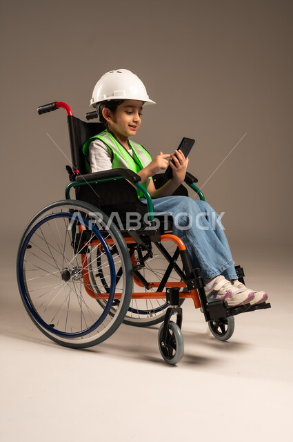 Browsing social media, using technical devices and modern technology, side portrait of a Saudi Arabian Gulf boy with special needs wearing a hat and a protective vest sitting in a wheelchair holding a mobile phone in his hand with gestures of pleasure, full body length, beige background