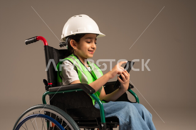 Browsing social media, using technical devices and modern technology, close-up side portrait of a Saudi Arabian Gulf boy with special needs wearing a hat and a protective vest sitting in a wheelchair holding a mobile phone in his hand and looking at it with gestures of pleasure, beige background