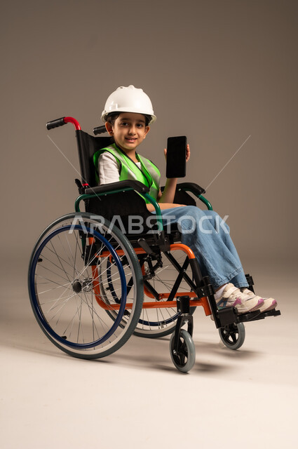 تجسيد العمل في المجال الهندسي منذ الصغر، عرض شاشة هاتف محمول سوداء فارغة، بورتريه من الجانب لولد عربي خليجي سعودي من ذوي الاحتياجات الخاصة يرتدي قبعة وسترة الحماية يجلس على كرسي متحرك يمسك الجوال وينظر للكاميرا بإيماءات السرور، الجسم بالطول الكامل، خلفية بيج