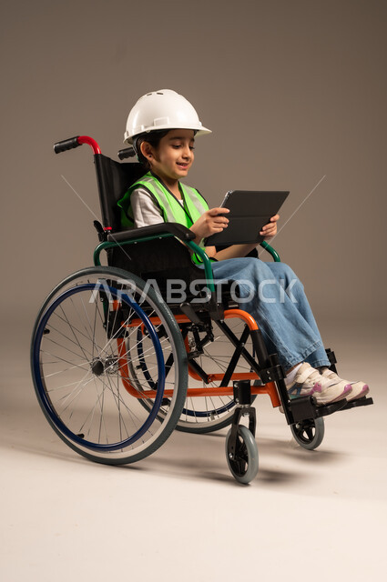 استخدام الأجهزة والتكنولوجيا الحديثة، التسلية واللعب الالكتروني، بورتريه من الجانب لولد عربي خليجي سعودي من ذوي الاحتياجات الخاصة يرتدي قبعة وسترة الحماية يجلس على كرسي متحرك يمسك جهاز التابلت بيده بإيماءات الاندماج والسرور، الجسم بالطول الكامل، خلفية بيج