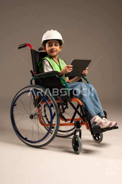 استخدام الأجهزة والتكنولوجيا الحديثة، التسلية واللعب الالكتروني، بورتريه من الجانب لولد عربي خليجي سعودي من ذوي الاحتياجات الخاصة يرتدي قبعة وسترة الحماية يجلس على كرسي متحرك يمسك جهاز التابلت بيده بإيماءات الاندماج والسرور، الجسم بالطول الكامل، خلفية بيج