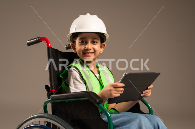 التسلية واللعب الالكتروني، استخدام الأجهزة التقنية والتكنولوجيا الحديثة، بورتريه مقرب من الجانب لولد عربي خليجي سعودي من ذوي الاحتياجات الخاصة يرتدي قبعة وسترة الحماية يجلس على كرسي متحرك يمسك التابلت بيده وينظر إلى الكاميرا بإيماءات السرور والبشاشة، خلفية بيج