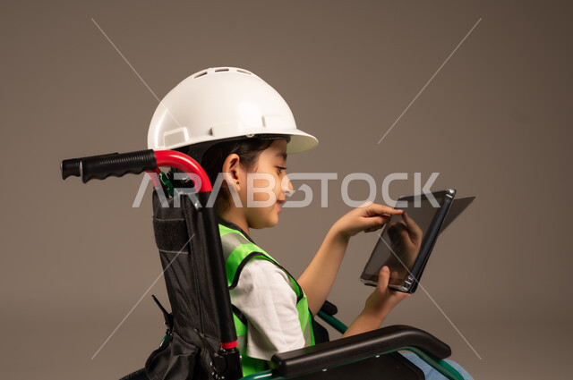 التسلية وقضاء وقت الفراغ باستخدام الأجهزة التقنية، دمج التكنولوجيا الحديثة بالحياة اليومية، بورتريه مقرب لولد عربي خليجي سعودي من ذوي الاحتياجات الخاصة يرتدي قبعة وسترة الحماية يجلس على كرسي متحرك يمسك جهاز التابلت بيده بإيماءات السرور، خلفية بيج