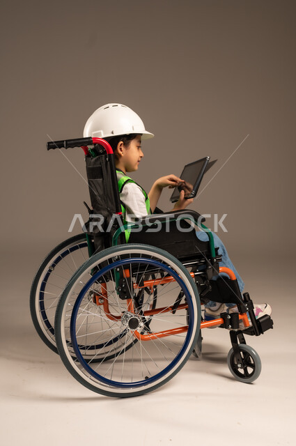 التسلية وقضاء وقت الفراغ باستخدام الأجهزة التقنية، دمج التكنولوجيا الحديثة بالحياة اليومية، بورتريه لولد عربي خليجي سعودي من ذوي الاحتياجات الخاصة يرتدي قبعة وسترة الحماية يجلس على كرسي متحرك يمسك جهاز التابلت بإيماءات السرور، الجسم بالطول الكامل، خلفية بيج