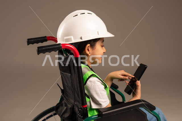 دمج التكنولوجيا الحديثة بالحياة اليومية، قضاء وقت الفراغ باستخدام الأجهزة التقنية، بورتريه مقرب من الجانب لولد عربي خليجي سعودي من ذوي الاحتياجات الخاصة يرتدي قبعة وسترة الحماية يجلس على كرسي متحرك يمسك جهاز الجوال بإيماءات الاندماج، خلفية بيج