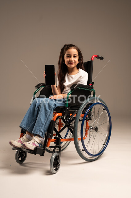 عرض شاشة هاتف محمول سوداء فارغة، طرق الإعلانات التسويقية المختلفة، بورتريه لفتاة صغيرة عربية خليجية سعودية من ذوي الاحتياجات الخاصة ترتدي الزي الكاجوال تجلس على كرسي متحرك تمسك جهاز الجوال بيدها وتنظر للكاميرا بإيماءات السرور، الجسم بالطول الكامل، خلفية بيج