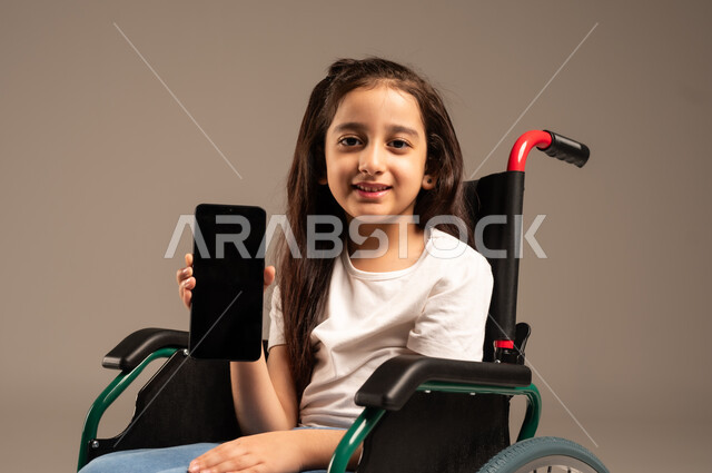 عرض شاشة هاتف محمول سوداء فارغة، طرق الإعلانات التسويقية المختلفة، بورتريه مقرب لفتاة صغيرة عربية خليجية سعودية من ذوي الاحتياجات الخاصة ترتدي الزي الكاجوال تجلس على كرسي متحرك تمسك جهاز الجوال بيدها وتنظر إلى الكاميرا بإيماءات السرور، خلفية بيج