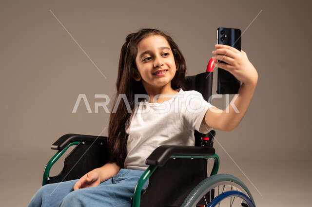 التقاط صور السيلفي التذكارية باستخدام الهاتف المحمول، بورتريه مقرب لفتاة صغيرة عربية خليجية سعودية من ذوي الاحتياجات الخاصة ترتدي الزي الكاجوال تجلس على كرسي متحرك تمسك جهاز الجوال بيدها وتأخذ لقطات عفوية بإيماءات السعادة، خلفية بيج