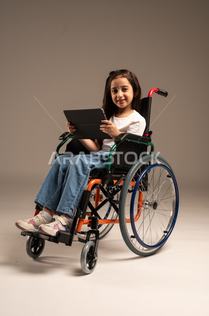 تصفح مواقع وتطبيقات الانترنت، دمج التكنولوجيا والأجهزة الحديثة بالحياة اليومية، بورتريه لفتاة صغيرة عربية خليجية سعودية من ذوي الاحتياجات الخاصة ترتدي الزي الكاجوال تجلس على كرسي متحرك تمسك جهاز التابلت بيدها بإيماءات السعادة، الجسم بالطول الكامل، خلفية بيج