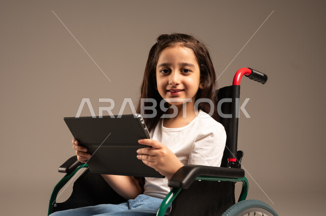 Integrating technology and modern devices into daily life, browsing websites and applications, close-up portrait of a young Saudi Arabian Gulf girl with special needs wearing casual clothes sitting on a wheelchair holding a tablet in her hand with gestures of happiness and pleasure, beige background