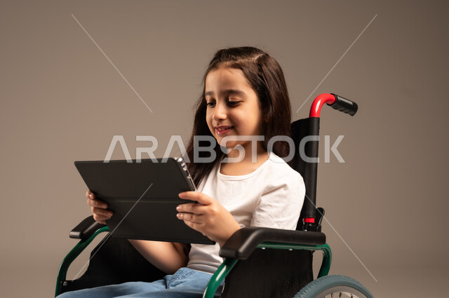 دمج التكنولوجيا والأجهزة الحديثة بالحياة اليومية، تصفح مواقع وتطبيقات الانترنت، بورتريه مقرب لفتاة صغيرة عربية خليجية سعودية من ذوي الاحتياجات الخاصة ترتدي الزي الكاجوال تجلس على كرسي متحرك تمسك جهاز التابلت بيدها بإيماءات السعادة والسرور، خلفية رمادية