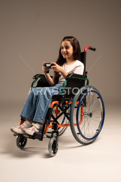 استخدام التقنيات والتكنولوجيا الحديثة، بورتريه لفتاة صغيرة عربية خليجية سعودية من ذوي الاحتياجات الخاصة ترتدي الزي الكاجوال تجلس على كرسي متحرك تمسك عصا التحكم بيدها بإيماءات الاندماج والتفاعل، الاستمتاع بألعاب الفيديو في المنزل، الجسم بالطول الكامل، خلفية بيج