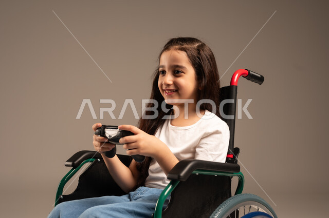  الاستمتاع بألعاب الفيديو في المنزل، استخدام التقنيات والتكنولوجيا الحديثة، بورتريه مقرب لفتاة صغيرة عربية خليجية سعودية من ذوي الاحتياجات الخاصة ترتدي الزي الكاجوال تجلس على كرسي متحرك تمسك عصا التحكم بيدها بإيماءات الاندماج والتفاعل، خلفية بيج