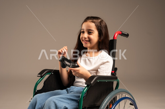  الاستمتاع بألعاب الفيديو في المنزل، استخدام التقنيات والتكنولوجيا الحديثة، بورتريه مقرب لفتاة صغيرة عربية خليجية سعودية من ذوي الاحتياجات الخاصة ترتدي الزي الكاجوال تجلس على كرسي متحرك تمسك عصا التحكم بيدها بإيماءات الاندماج والتفاعل، خلفية بيج