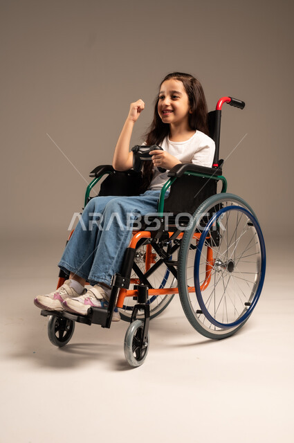 استخدام التقنيات والتكنولوجيا الحديثة، بورتريه لفتاة صغيرة عربية خليجية سعودية من ذوي الاحتياجات الخاصة ترتدي الزي الكاجوال تجلس على كرسي متحرك تمسك عصا التحكم بيدها بإيماءات الاندماج والتفاعل، الاستمتاع بألعاب الفيديو في المنزل، الجسم بالطول الكامل، خلفية بيج