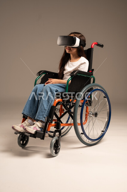 Using virtual reality techniques and modern technology, portrait of a young Saudi Arabian Gulf girl with special needs wearing casual clothes sitting in a wheelchair wearing VR glasses, poor motor ability and difficulty walking, full body, beige background