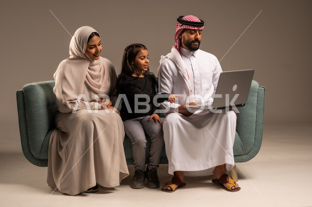 مشاهدة الافلام والبرامج الترفيهيه، القيام بالانشطة المسلية في المنزل، قضاء وقت ممتع مع الأبناء، بورتريه لرجل عربي خليجي سعودي يرتدي الثوب التقليدي والشماع يجلس على الكنبة بجانب ابنته وزوجته يقومون باجراء مكالمة فيديو عبر الحاسوب المحمول، خلفية رمادية