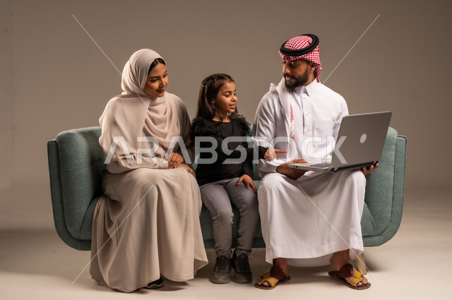 مشاهدة الافلام والبرامج الترفيهيه، القيام بالانشطة المسلية في المنزل، قضاء وقت ممتع مع الأبناء، بورتريه لرجل عربي خليجي سعودي يرتدي الثوب التقليدي والشماع يجلس على الكنبة بجانب ابنته وزوجته يقومون باجراء مكالمة فيديو عبر الحاسوب المحمول، خلفية رمادية