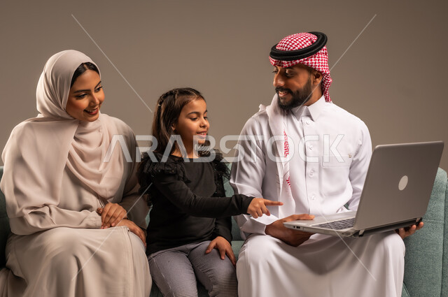 مشاهدة الافلام والبرامج الترفيهيه، القيام بالانشطة المسلية في المنزل، قضاء وقت ممتع مع الأبناء، بورتريه لرجل عربي خليجي سعودي يرتدي الثوب التقليدي والشماع يجلس على الكنبة بجانب ابنته وزوجته يقومون باجراء مكالمة فيديو عبر الحاسوب المحمول، خلفية رمادية