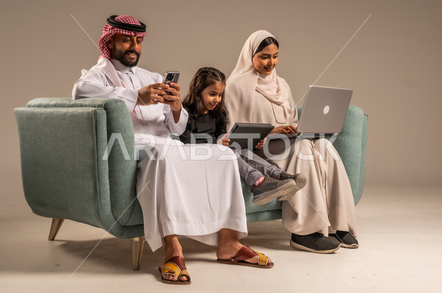 مشاهدة الافلام والبرامج الترفيهيه، القيام بالانشطة المسلية في المنزل، قضاء وقت ممتع مع الأبناء، بورتريه لرجل عربي خليجي سعودي يرتدي الثوب التقليدي والشماع يجلس على الكنبة بجانب ابنته وزوجته يقومون باجراء مكالمة فيديو عبر الحاسوب المحمول، خلفية رمادية