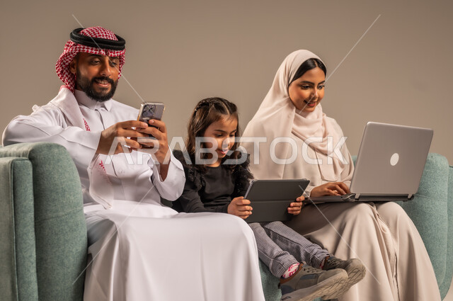 مشاهدة الافلام والبرامج الترفيهيه، القيام بالانشطة المسلية في المنزل، قضاء وقت ممتع مع الأبناء، بورتريه لرجل عربي خليجي سعودي يرتدي الثوب التقليدي والشماع يجلس على الكنبة بجانب ابنته وزوجته يقومون باجراء مكالمة فيديو عبر الحاسوب المحمول، خلفية رمادية