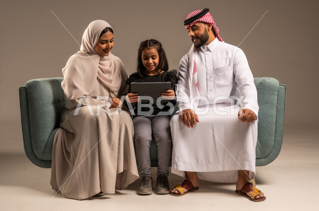 قضاء وقت ممتع مع الأبناء، مشاهدة الافلام والبرامج الترفيهيه، القيام بالانشطة المسلية في المنزل،  بورتريه لرجل عربي خليجي سعودي يرتدي الثوب التقليدي والشماع يجلس على الكنبة بجانب ابنته وزوجته يقومون باجراء مكالمة فيديو عبر الجهاز اللوحي، خلفية رمادية