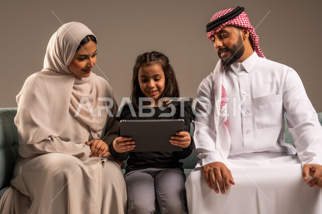قضاء وقت ممتع مع الأبناء، مشاهدة الافلام والبرامج الترفيهيه، القيام بالانشطة المسلية في المنزل،  بورتريه لرجل عربي خليجي سعودي يرتدي الثوب التقليدي والشماع يجلس على الكنبة بجانب ابنته وزوجته يقومون باجراء مكالمة فيديو عبر الجهاز اللوحي، خلفية رمادية