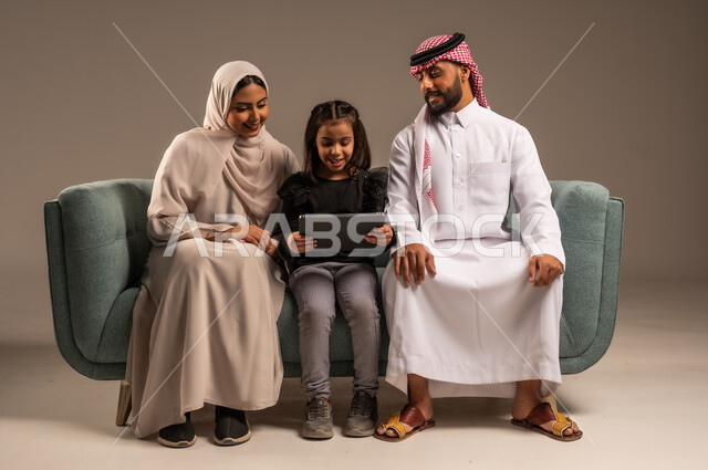 قضاء وقت ممتع مع الأبناء، مشاهدة الافلام والبرامج الترفيهيه، القيام بالانشطة المسلية في المنزل،  بورتريه لرجل عربي خليجي سعودي يرتدي الثوب التقليدي والشماع يجلس على الكنبة بجانب ابنته وزوجته يقومون باجراء مكالمة فيديو عبر الجهاز اللوحي، خلفية رمادية