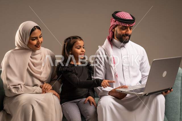 القيام بالانشطة المسلية في المنزل، قضاء وقت ممتع مع الأبناء، مشاهدة الافلام والبرامج الترفيهيه،  بورتريه لرجل عربي خليجي سعودي يرتدي الثوب التقليدي والشماع يجلس على الكنبة بجانب ابنته وزوجته يقومون باجراء مكالمة فيديو عبر الحاسوب المحمول، خلفية رمادية