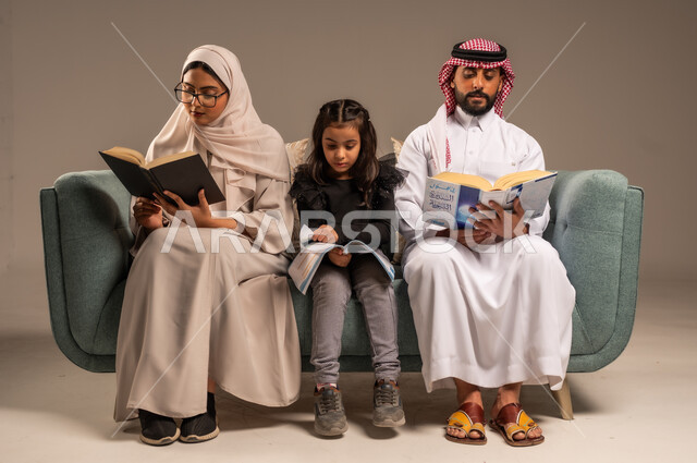 شغف المطالعة للكتب والروايات، القيام بالانشطة المسلية في المنزل، تعليم الاولاد على التثقيف والمطالعة، قضاء وقت ممتع مع الأبناء، بورتريه لرجل عربي خليجي سعودي يجلس على الكنبة  بجانب زوجته وابنته يقومون بقراءة القصص، تنمية المهارات الأدبية والفنية، خلفية رمادية