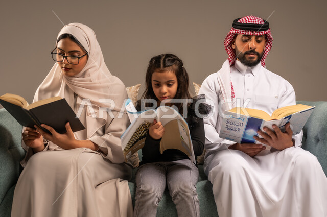 شغف المطالعة للكتب والروايات، القيام بالانشطة المسلية في المنزل، تعليم الاولاد على التثقيف والمطالعة، قضاء وقت ممتع مع الأبناء، بورتريه لرجل عربي خليجي سعودي يجلس على الكنبة  بجانب زوجته وابنته يقومون بقراءة القصص، تنمية المهارات الأدبية والفنية، خلفية رمادية