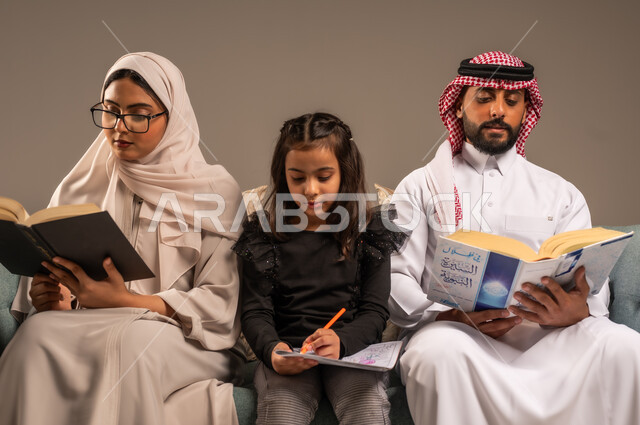 شغف المطالعة للكتب والروايات، القيام بالانشطة المسلية في المنزل، تعليم الاولاد على التثقيف والمطالعة، قضاء وقت ممتع مع الأبناء، بورتريه لرجل عربي خليجي سعودي يجلس على الكنبة  بجانب زوجته وابنته يقومون بقراءة القصص، تنمية المهارات الأدبية والفنية، خلفية رمادية