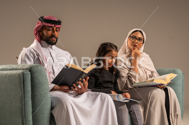  تنمية المهارات الأدبية والفنية، شغف المطالعة للكتب والروايات، القيام بالانشطة المسلية في المنزل، تعليم الاولاد على التثقيف والمطالعة، قضاء وقت ممتع مع الأبناء، بورتريه لرجل عربي خليجي سعودي يجلس على الكنبة  بجانب زوجته وابنته يقومون بقراءة القصص، خلفية رمادية