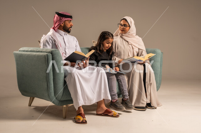 تنمية المهارات الأدبية والفنية، شغف المطالعة للكتب والروايات، القيام بالانشطة المسلية في المنزل، تعليم الاولاد على التثقيف والمطالعة، قضاء وقت ممتع مع الأبناء، بورتريه لرجل عربي خليجي سعودي يجلس على الكنبة  بجانب زوجته وابنته يقومون بقراءة القصص، خلفية رمادية