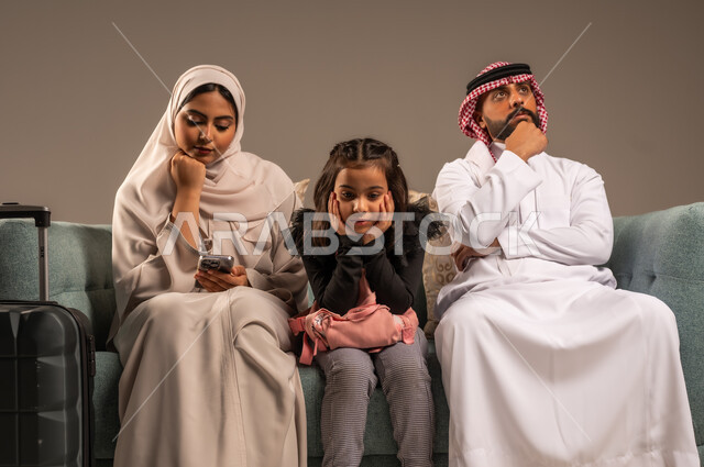انتظار موعد الطائرة بايماءات الملل، بورتريه لرجل عربي خليجي سعودي يرتدي الثوب التقليدي والشماغ يجلس على كرسي مريح مع زوجته وابنته يمسك حقيبة الأمتعة، الإستعداد لقضاء الإجازة الصيفية والذهاب في رحلة سفر ممتعة، الأجواء العائلية المليئة بالحب،  خلفية رمادية