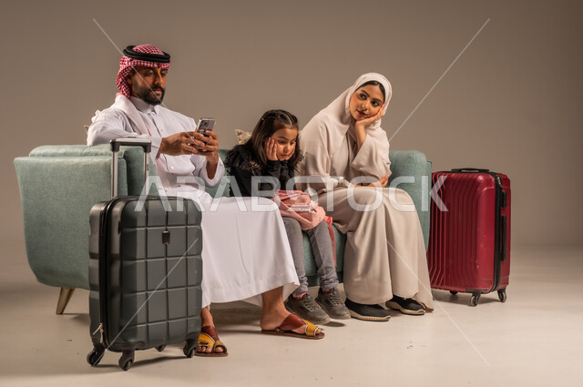 انتظار موعد الطائرة بايماءات الملل، بورتريه لرجل عربي خليجي سعودي يرتدي الثوب التقليدي والشماغ يجلس على كرسي مريح مع زوجته وابنته يمسك حقيبة الأمتعة، الإستعداد لقضاء الإجازة الصيفية والذهاب في رحلة سفر ممتعة، الأجواء العائلية المليئة بالحب،  خلفية رمادية